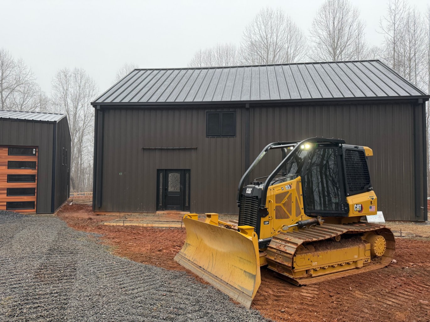 A yellow bulldozer is parked on red soil in front of a dark metal building with a garage on a cloudy day; bare trees are in the background.
