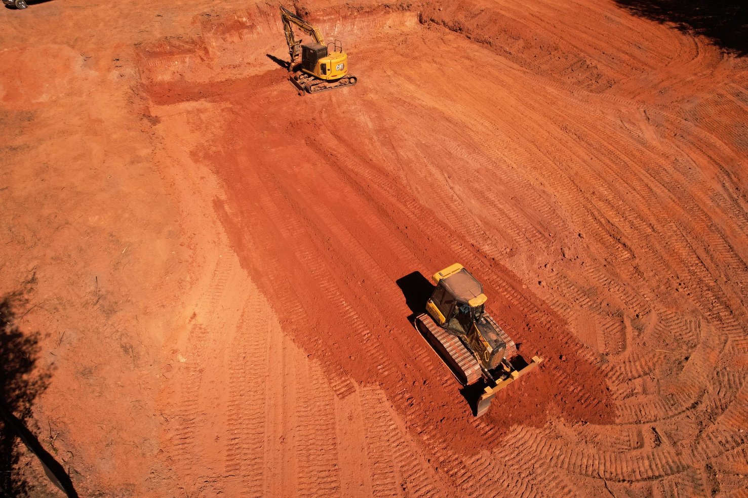 Two construction vehicles operate on a large, cleared area of red earth, with visible tire and track marks covering the ground.