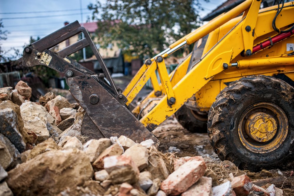 A yellow backhoe loader moves rubble and rocks at a construction or demolition site, with buildings and trees in the background.