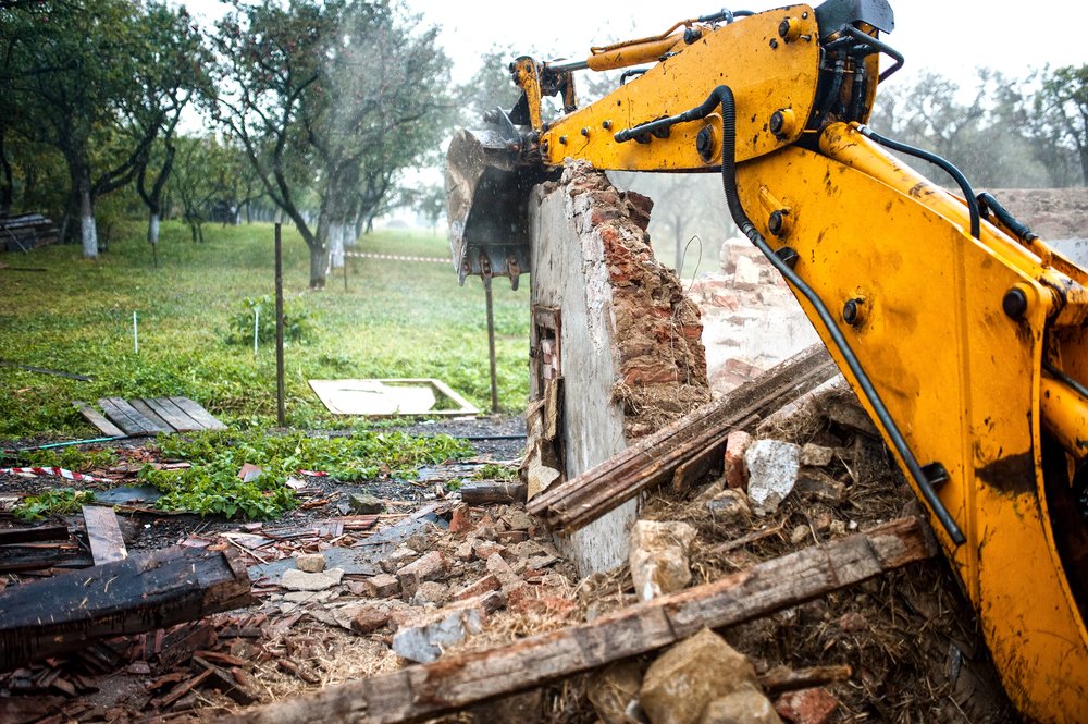 A yellow excavator demolishes a brick wall outdoors, with debris scattered on the ground and trees in the background.