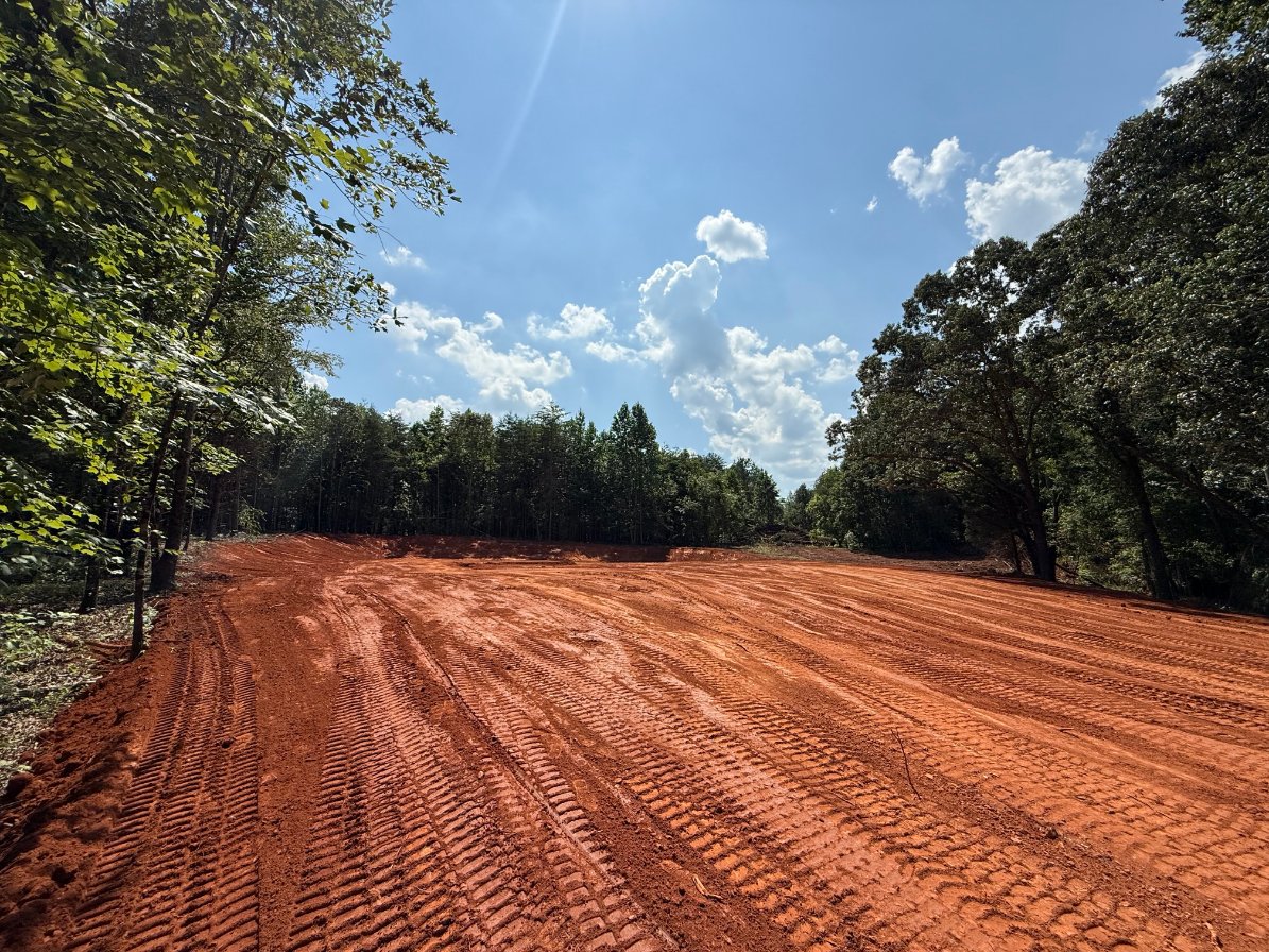 A freshly graded red dirt field with visible tire tracks, surrounded by trees under a partly cloudy blue sky.