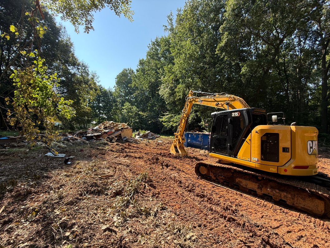 A yellow excavator sits on a cleared dirt lot next to a pile of debris and fallen trees, surrounded by wooded area under a clear sky.