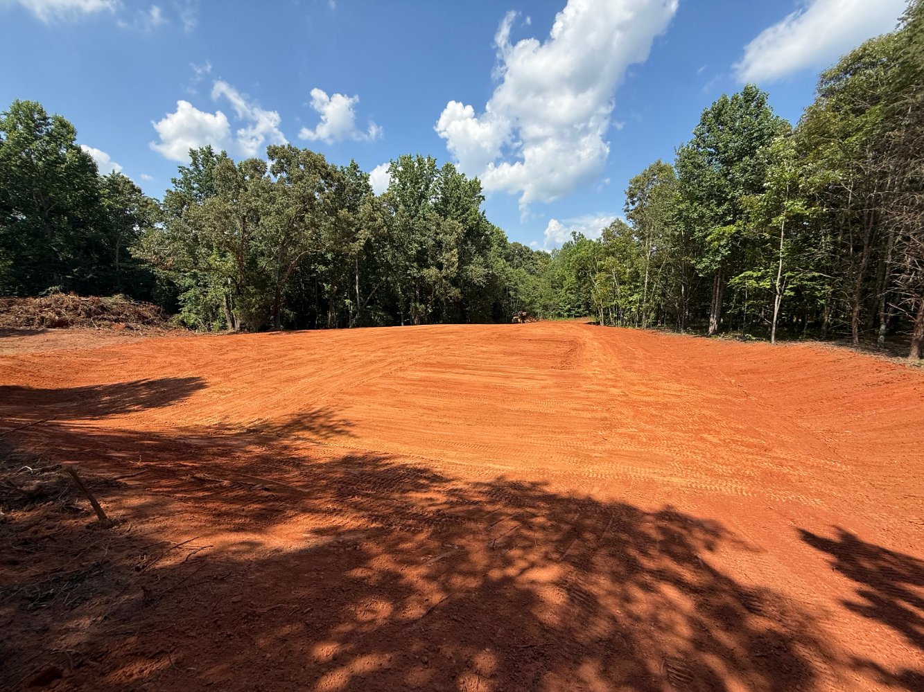 A large, freshly graded area of red dirt surrounded by green trees under a partly cloudy blue sky.