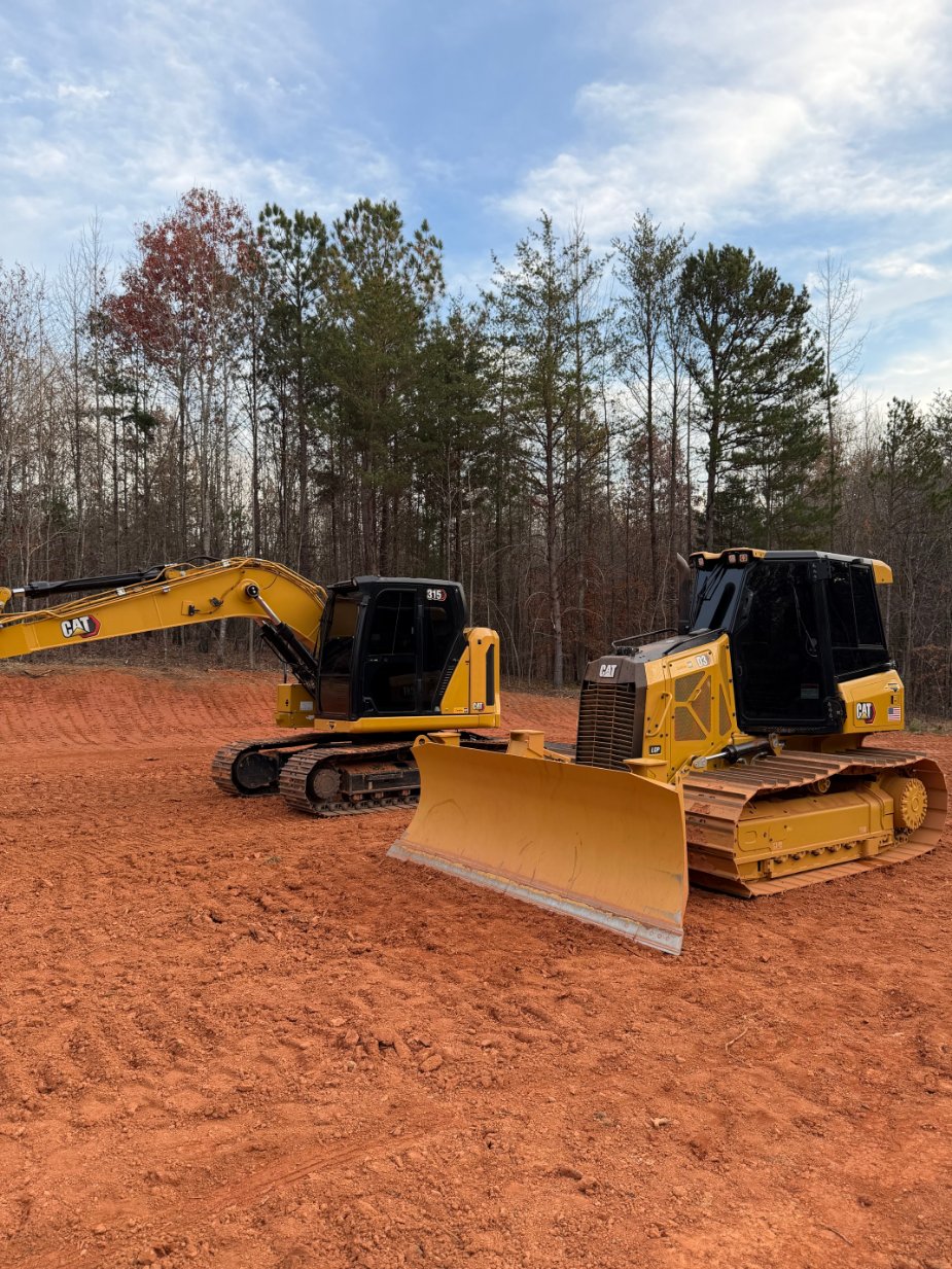 A yellow excavator and a yellow bulldozer are parked on a cleared, red dirt area with trees in the background under a partly cloudy sky.