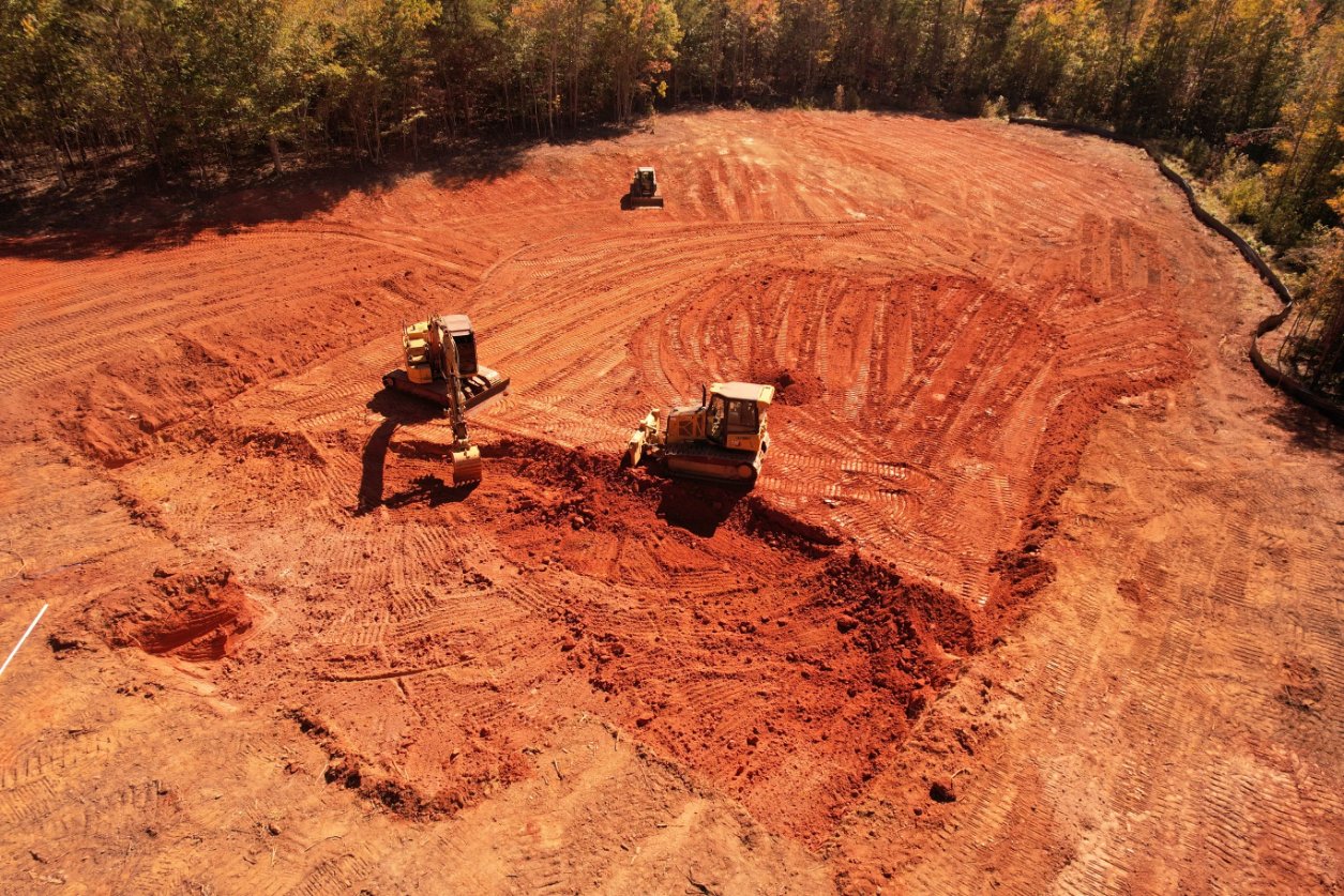 Two construction vehicles, a bulldozer and an excavator, are clearing and leveling red soil on a large, open site surrounded by trees.