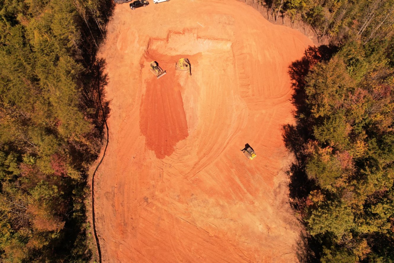 Aerial view of a construction site with red soil, surrounded by trees, showing three pieces of heavy machinery working on the cleared land.