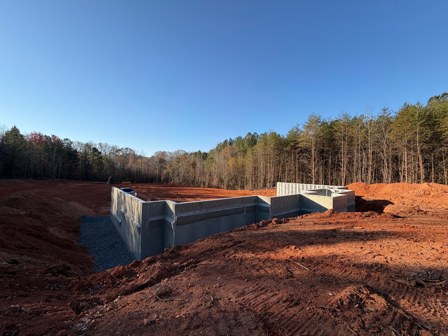 A concrete foundation of a building under construction is set in a cleared, red soil area surrounded by trees under a clear blue sky.
