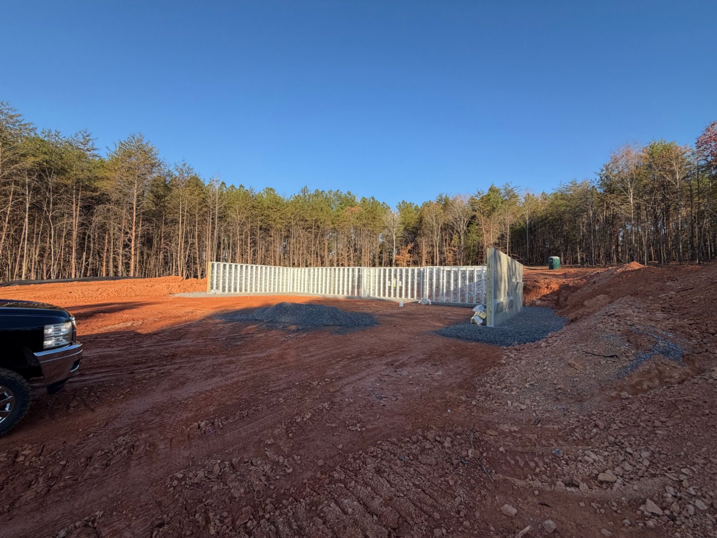 Construction site with newly built concrete foundation walls surrounded by red dirt, gravel, and forested area under a clear blue sky.